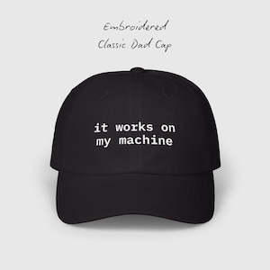 May include: Black classic dad cap with the embroidered text "it works on my machine" in white. The cap also has the text "Embroidered Classic Dad Cap" in cursive. The hat is a baseball style cap.