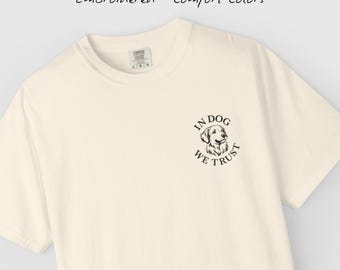 May include: Cream-colored t-shirt with the phrase "In Dog We Trust" and a dog illustration. The shirt also has the words "Embroidered • Comfort Colors" printed on it.
