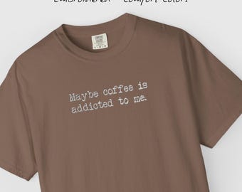 May include: A brown t-shirt with the embroidered text "Maybe coffee is addicted to me." The shirt also has the text "Embroidered • Comfort Colors" at the top. The shirt has a crew neck and short sleeves.