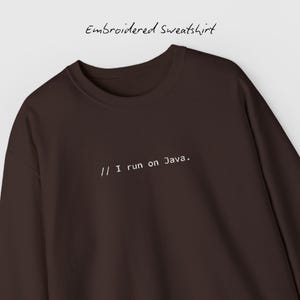 Op de afbeelding: Een donkerbruine geborduurde sweatshirt met de tekst "// I run on Java." in het wit. De woorden "Embroidered Sweatshirt" staan erboven. De sweatshirt is effen van kleur en heeft een ronde hals.