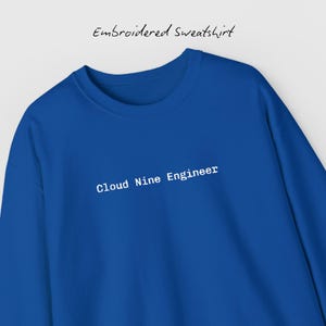 May include: A cobalt blue sweatshirt featuring the white text "Cloud Nine Engineer". The words "Embroidered Sweatshirt" are written above the text. The sweatshirt is a solid colour and has a crew neck.