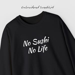 Op de afbeelding: Zwart geborduurd sweatshirt met de witte tekst "No Sushi No Life". De sweatshirt heeft een ronde hals en lange mouwen. De woorden "Embroidered Sweatshirt" staan boven de hoofdtekst.
