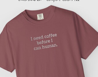 May include: A maroon-coloured t-shirt with the embroidered phrase, "I need coffee before I can human." The shirt also has a small tag that says "Comfort Colors". The shirt is a short-sleeved crew neck.