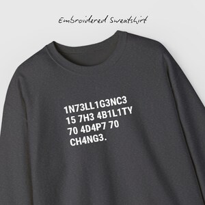 May include: Dark gray embroidered sweatshirt with white text that reads "1N73LL1G3NC3 15 7H3 4B1L1TY 70 4D4P7 70 CH4NG3." The text "Embroidered Sweatshirt" is above the main text.