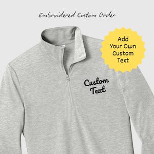 Peut inclure: Un pull-over gris clair à quart de fermeture éclair avec un col et une fermeture éclair. Le texte "Custom Text" est brodé sur le devant. Un graphique en forme d'éclat jaune indique "Add Your Own Custom Text". Les mots "Embroidered Custom Order" sont au-dessus.