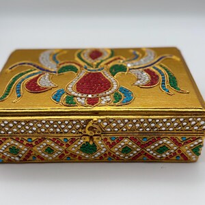 Op de afbeelding: Een rechthoekige, goudkleurige decoratieve doos met ingewikkelde, kleurrijke patronen. Het deksel heeft een centraal ontwerp in rood, wit en groen, met blauwe en witte accenten. De randen van de doos zijn versierd met een herhalend patroon van wit, rood, groen en blauw.
