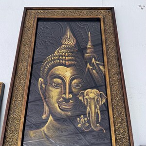 May include: A framed artwork depicting a golden Buddha and an elephant against a dark backdrop. The frame has an elaborate, gold-coloured design. This is likely a religious or decorative item, suitable for display.