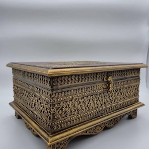 May include: Ornate, rectangular box in shades of gold and brown. The box features intricate, repeating patterns and rests on four decorative feet. The lid is slightly raised, and a small clasp is visible on the front. The box is likely used for storage.