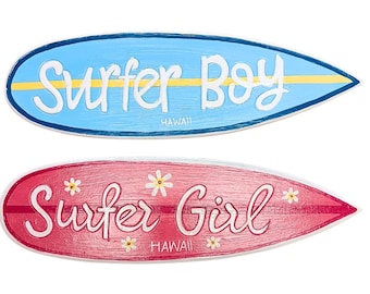 May include: Two surfboard-shaped wooden signs. One is light blue with "Surfer Boy" in white script and a yellow stripe. The other is red with "Surfer Girl" in white script and small flower accents. Both say "HAWAII".