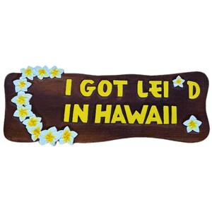 May include: A wooden sign with the phrase "I GOT LEI'D IN HAWAII" in yellow letters. The sign is decorated with white and yellow flower accents, evoking a tropical theme.