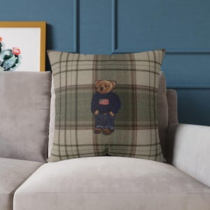 Puede incluir: Un cojín cuadrado con un patrón de cuadros en tonos beige y verde oliva. En el centro del cojín hay un gráfico de un oso de peluche marrón que viste un suéter azul marino con una bandera estadounidense, pantalones vaqueros y zapatos marrones.