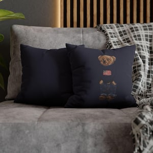 Puede incluir: Dos cojines cuadrados de color azul marino oscuro. El cojín de la derecha presenta un gráfico central de un oso de peluche marrón que lleva un suéter oscuro con una pequeña bandera estadounidense, vaqueros azules y zapatos marrones, con las manos en los bolsillos.