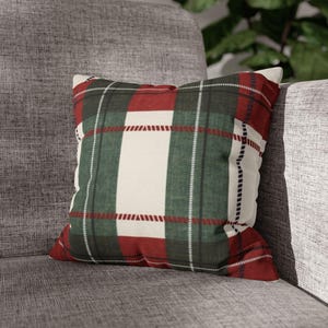 Puede incluir: Un cojín decorativo cuadrado con un estampado de cuadros en rojo, verde y blanco. El cojín está colocado sobre un sofá gris tapizado. El diseño de cuadros presenta líneas gruesas y finas, creando un aspecto clásico y acogedor. Es un artículo de decoración del hogar.