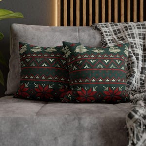 Puede incluir: Dos cojines decorativos con un fondo verde oscuro y un patrón repetido de copos de nieve y formas geométricas rojas y crema. Los cojines están colocados sobre un sofá gris, lo que sugiere un tema de diseño de interiores acogedor.