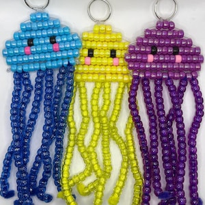 May include: Three jellyfish-shaped keychains made from plastic pony beads. The left keychain is light blue with dark blue tentacles, the center is yellow with yellow tentacles, and the right is purple with purple tentacles. Each has a metal ring.