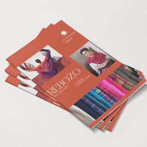 May include: A stack of brochures with an orange cover featuring the word "REBOZO" and images of textiles and people wearing shawls. The brochures are titled "A Thread Through Time" and "By Sara Luna 2025".