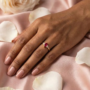 May include: A gold ring featuring a marquise-cut ruby gemstone, flanked by smaller white gemstones. The ring is set on a hand with a pink manicure, against a backdrop of pink fabric and white flower petals.