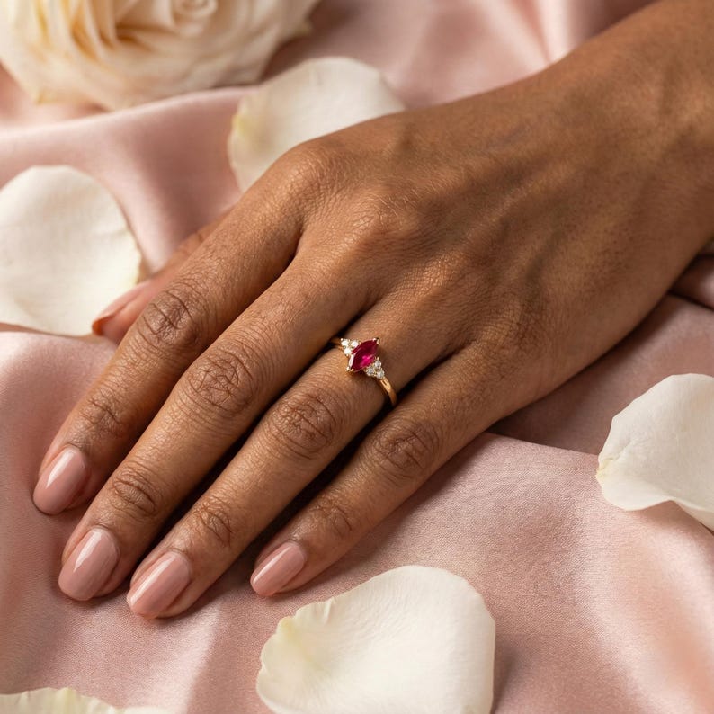 May include: A gold ring featuring a marquise-cut ruby gemstone, flanked by smaller white gemstones. The ring is set on a hand with a pink manicure, against a backdrop of pink fabric and white flower petals.