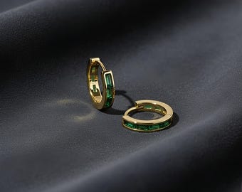 May include: A pair of gold hoop earrings with emerald green rectangular stones. The earrings are set against a dark background, highlighting their bright color and design. The jewelry is a classic accessory.