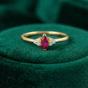 May include: A gold ring featuring a marquise-cut ruby gemstone, flanked by small, round, clear gemstones. The ring's band is a simple, polished gold. The ruby is a deep, vibrant red, and the ring is set against a dark green background.