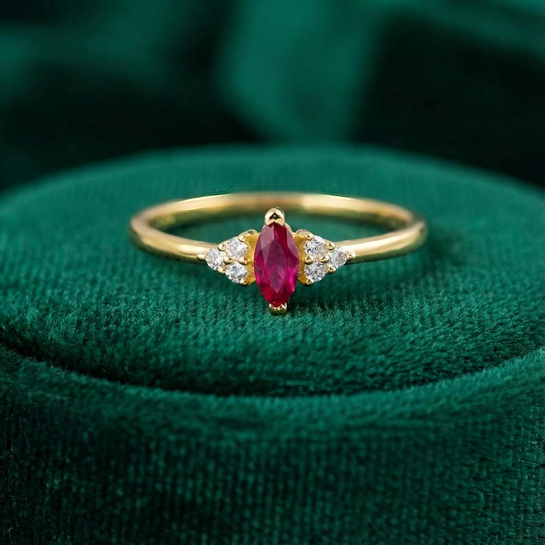 May include: A gold ring featuring a marquise-cut ruby gemstone, flanked by small, round, clear gemstones. The ring's band is a simple, polished gold. The ruby is a deep, vibrant red, and the ring is set against a dark green background.