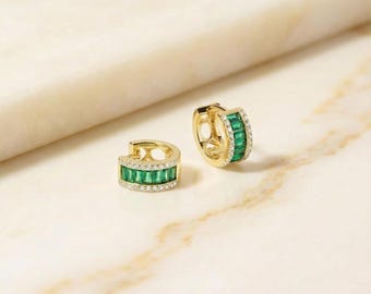 May include: A pair of gold hoop earrings with emerald green and clear stones. The earrings have a rectangular emerald green stone design in the center and clear stones around the edges. The earrings are displayed on a white and tan marble surface.