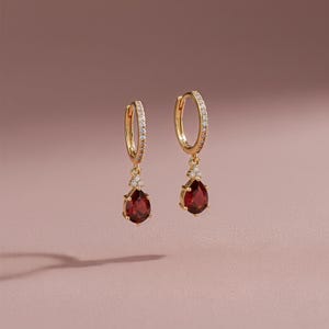 以下が含まれることがあります： ゴールドカラーのフープピアス2つ。涙型の深い赤色の宝石ペンダントが付いています。フープには小さな透明な宝石がちりばめられています。ピアスはソフトピンクの背景に表示されています。