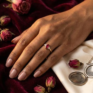 May include: A gold ring with a marquise-cut red gemstone and small white accent stones. The ring is on a hand with manicured nails, set against a backdrop of red velvet, dried roses, and a vintage locket.