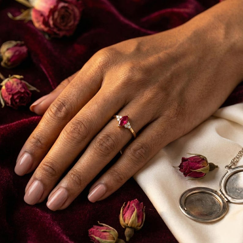 May include: A gold ring with a marquise-cut red gemstone and small white accent stones. The ring is on a hand with manicured nails, set against a backdrop of red velvet, dried roses, and a vintage locket.