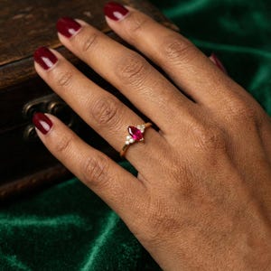 May include: A gold ring featuring a pear-shaped, deep red gemstone, accented with small, round white gemstones. The ring is set on a hand with dark skin, the nails painted a matching deep red. The background is a dark green velvet.