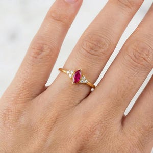 May include: A gold ring featuring a marquise-cut ruby center stone, flanked by small, round, clear gemstones. The ring is set on a thin, gold band. The ring is shown on a person's finger.