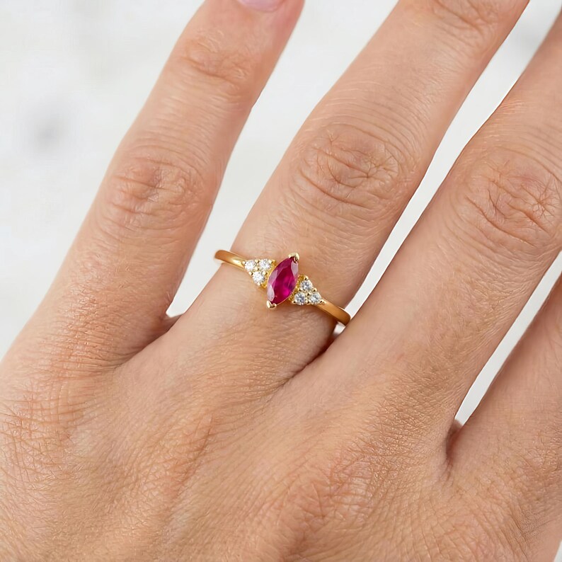 May include: A gold ring featuring a marquise-cut ruby center stone, flanked by small, round, clear gemstones. The ring is set on a thin, gold band. The ring is shown on a person's finger.