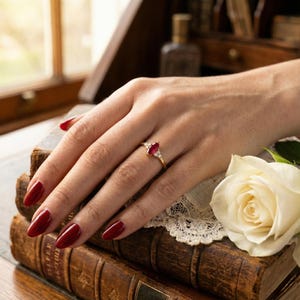 May include: A gold ring featuring a marquise-cut ruby gemstone, flanked by smaller white gemstones. The ring is set on a hand with red nail polish, resting on antique books and a white rose.