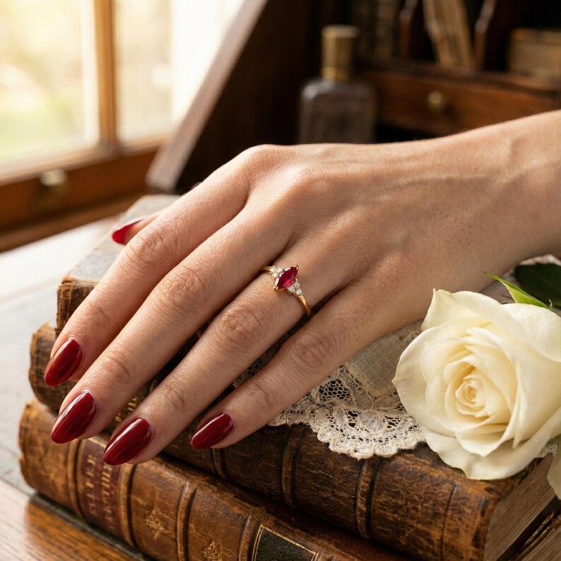 May include: A gold ring featuring a marquise-cut ruby gemstone, flanked by smaller white gemstones. The ring is set on a hand with red nail polish, resting on antique books and a white rose.
