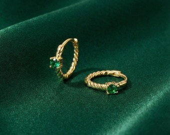 May include: Two gold-colored hoop earrings with a twisted rope design. Each earring features a square-cut, emerald-green gemstone. The earrings are displayed on a dark green, textured fabric.