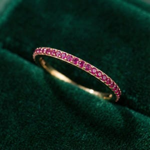 Pode incluir: Um anel de banda dourada com uma fileira contínua de pequenas e vibrantes pedras preciosas rosa. O anel é apresentado sobre um fundo de veludo verde escuro, realçando a cor e o design do anel. O anel é uma joia.