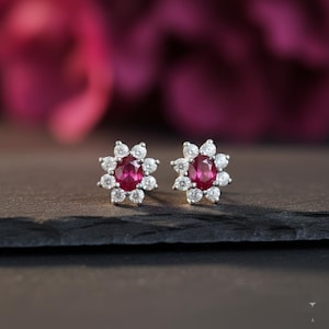 Pode incluir: Um par de brincos de botão em forma de flor. Cada brinco apresenta um rubi oval central rodeado por um halo de pequenas pedras brancas redondas. Os brincos estão colocados sobre uma superfície escura com um fundo desfocado rosa escuro.