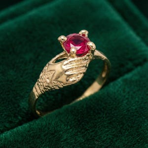 May include: A gold ring with a large, round, red gemstone. The ring's design includes a hand-shaped element holding the gemstone. The background is a dark green colour.