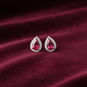 Puede incluir: Un par de pendientes en forma de lágrima con una vibrante gema rosa en el centro, rodeada por un halo de pequeñas piedras blancas brillantes. El fondo es de terciopelo burdeos oscuro, creando una sensación de lujo.