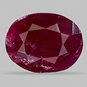 May include: A faceted, oval-shaped ruby gemstone. The deep red gem has a starburst pattern of light reflecting off its many facets. The ruby is set against a plain, light gray background.