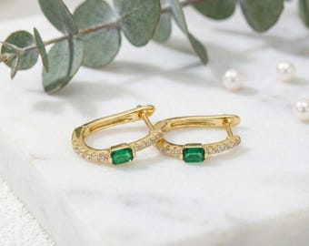 May include: A pair of gold hoop earrings with emerald-cut green gemstones. The earrings are encrusted with small, clear gemstones along the bottom curve. The earrings are displayed on a white marble surface with greenery and pearls in the background.