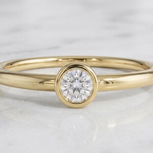 May include: A yellow gold ring featuring a single round brilliant-cut white diamond set in a smooth bezel setting. The band is polished and has a rounded profile, showcasing a clean and simple design with a high-shine metallic finish.