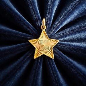 May include: A gold star pendant with a textured, sunburst pattern. The star hangs from a small loop, set against a dark blue, velvet fabric background.