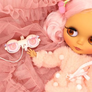 May include: A doll with pink hair and brown eyes, wearing a pink outfit, rests on a pink tulle fabric. A white and pink eye mechanism with a wire is next to the doll. The doll has a fair complexion and is adorned with pink accessories.