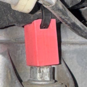 May include: Close-up of a red rectangular component attached to a metal bolt. The component is positioned near a bundle of wires and a black cable. The background is a muted gray, suggesting an automotive or mechanical setting.