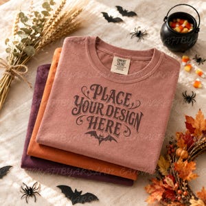 May include: A stack of three folded t-shirts in muted rust, orange, and dark purple colors. The top shirt features the text "PLACE YOUR DESIGN HERE" in dark brown lettering above a small bat graphic and a Comfort Colors brand neck label.