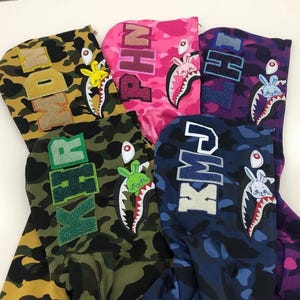 May include: A collection of camouflage hoodies in various colors, including yellow, pink, green, blue, and purple. Each hoodie features a shark mouth design on the hood and a bunny graphic. The hoods also display different letter combinations.