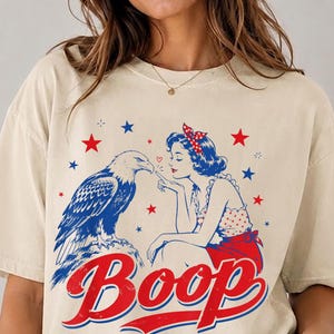 May include: A beige short-sleeve t-shirt featuring a graphic print of a bald eagle and a pin-up style figure with the word Boop in red and blue. The design includes red and blue stars and a polka dot pattern on the figure's top.