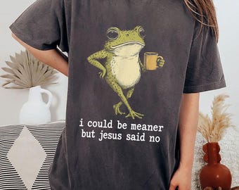 May include: Dark grey t-shirt with a cartoon frog holding a mug. The frog is green with yellow spots. The text "i could be meaner but jesus said no" is printed below the frog in white lettering.