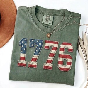 May include: An olive green t-shirt featuring the year "1776" in a patriotic design. The numbers are in red, white, and blue, resembling the American flag. A gold necklace with a pendant rests on the shirt.